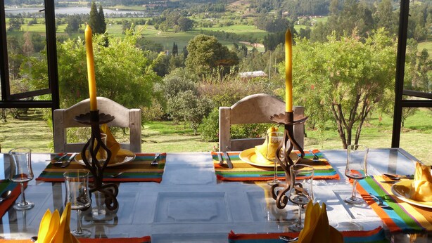 Una mesa de comedor para dos, con velas, copas y un mantel individual colorido, con vistas a un paisaje pintoresco.