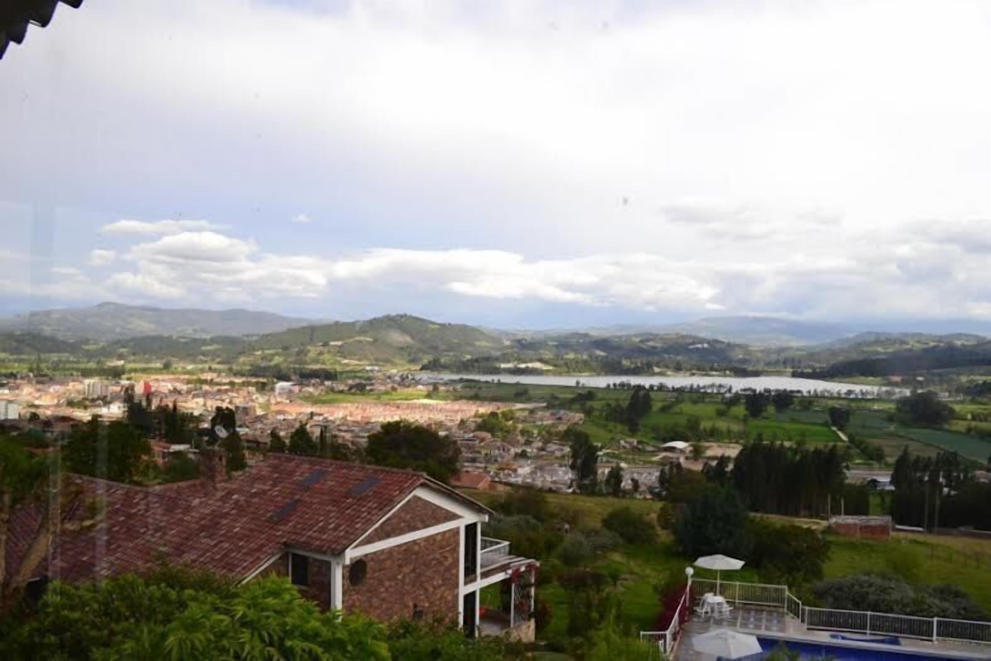 Vista de un pueblo con un edificio destacado al frente, una masa de agua a lo lejos y colinas onduladas en el fondo.