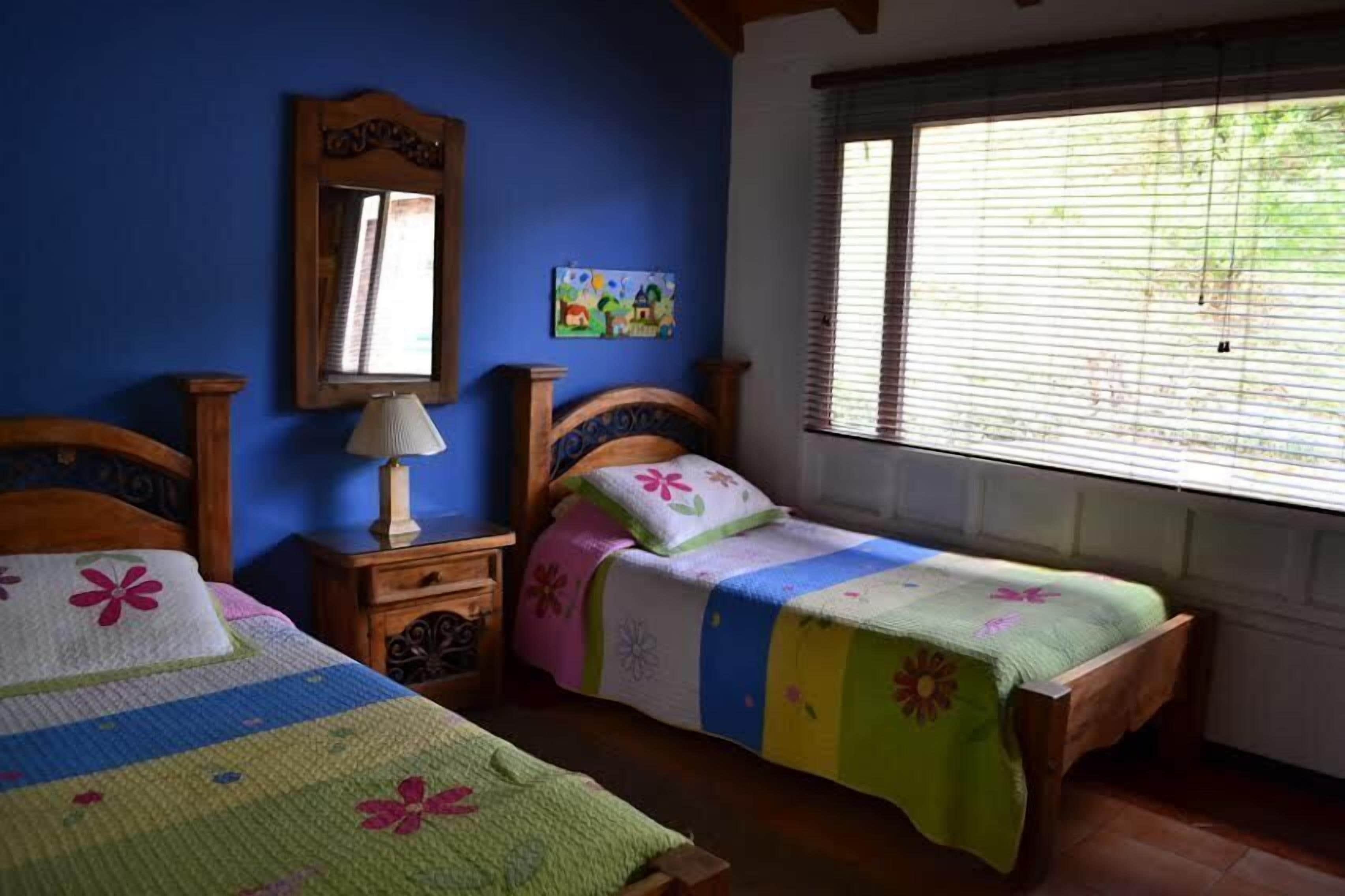 Een slaapkamer met twee houten bedden, een nachtkastje met een lamp, een spiegel en een raam met jaloezieën.
