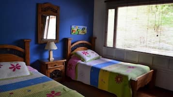 Een slaapkamer met twee houten bedden, een nachtkastje met een lamp, een spiegel en een raam met jaloezieën.