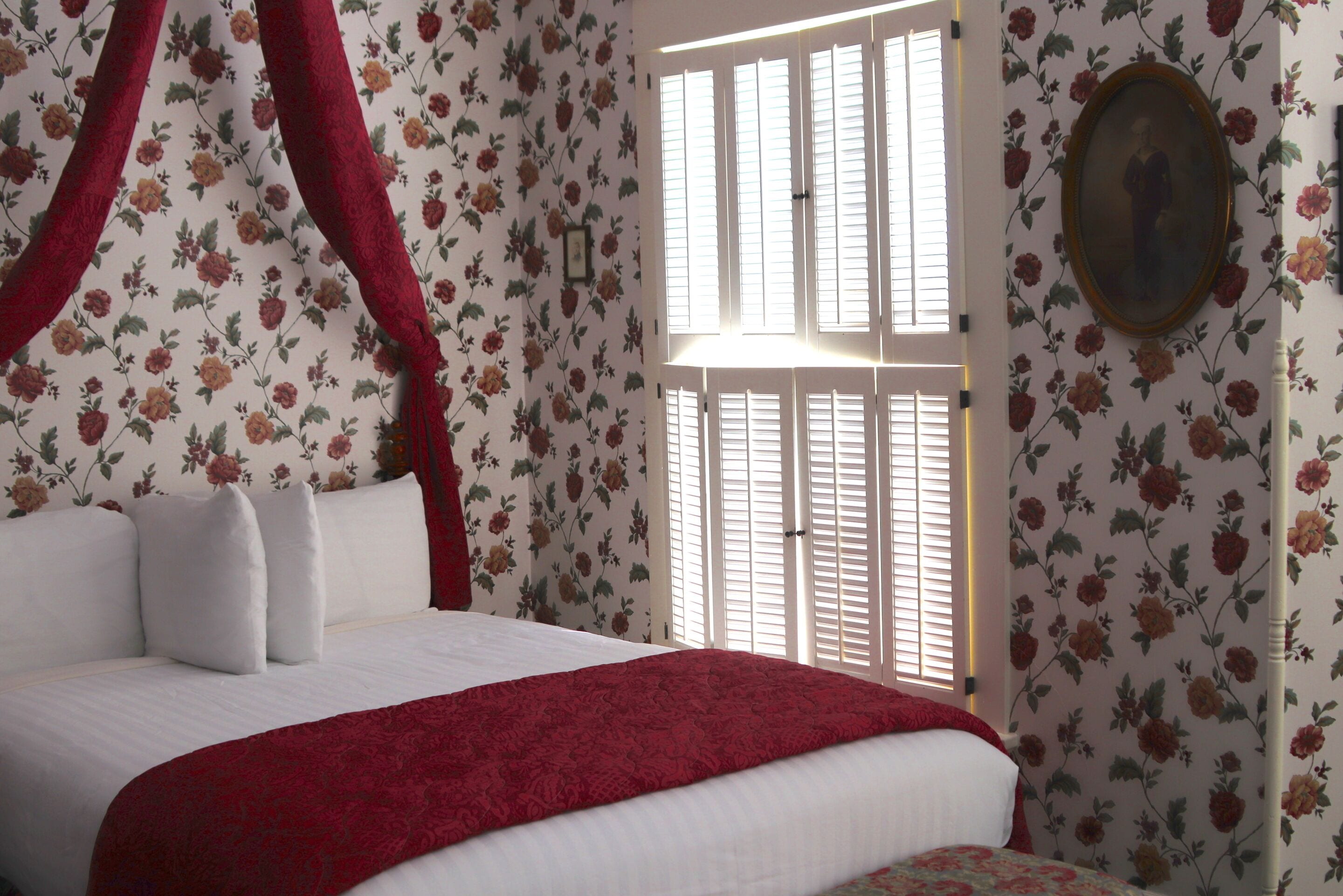 Ein Schlafzimmer mit Bett, blumigem Wandpapier, einem Fenster mit Fensterläden und einem gerahmten Bild an der Wand.