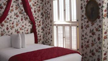 Ein Schlafzimmer mit Bett, blumigem Wandpapier, einem Fenster mit Fensterläden und einem gerahmten Bild an der Wand.