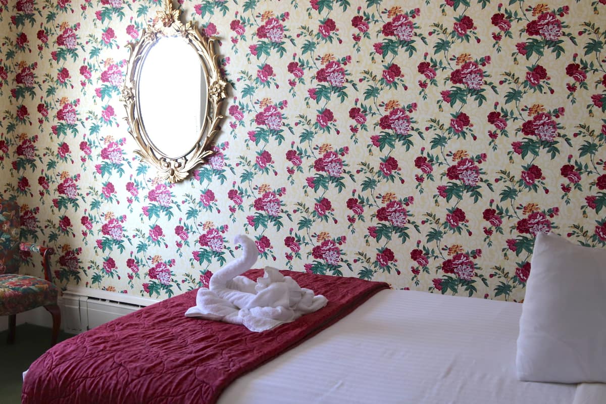 A hotel room with floral wallpaper, a bed with a red blanket, a swan-shaped towel, a white pillow, and a mirror with an ornate frame.