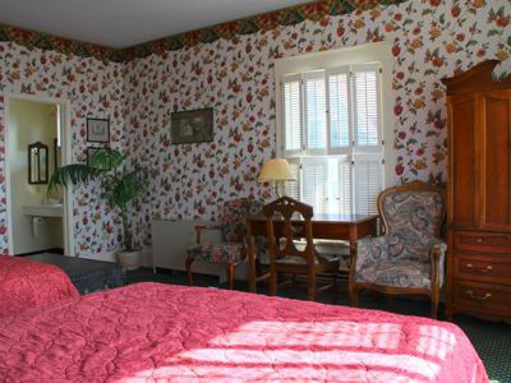 A room with floral wallpaper, a bed with a pink quilt, a wooden chair, a table, and a cabinet.