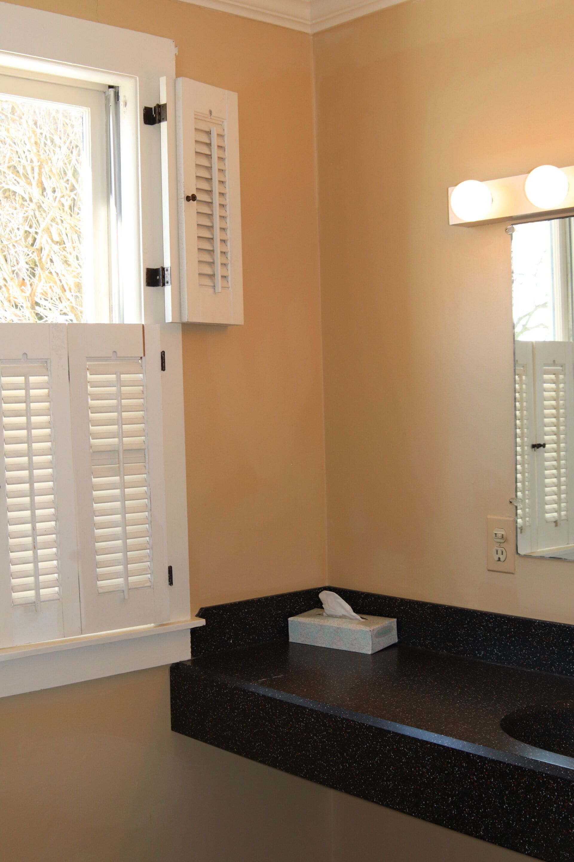A bathroom with a black granite countertop, a white door with shutters, and a mirror with lights above it.
