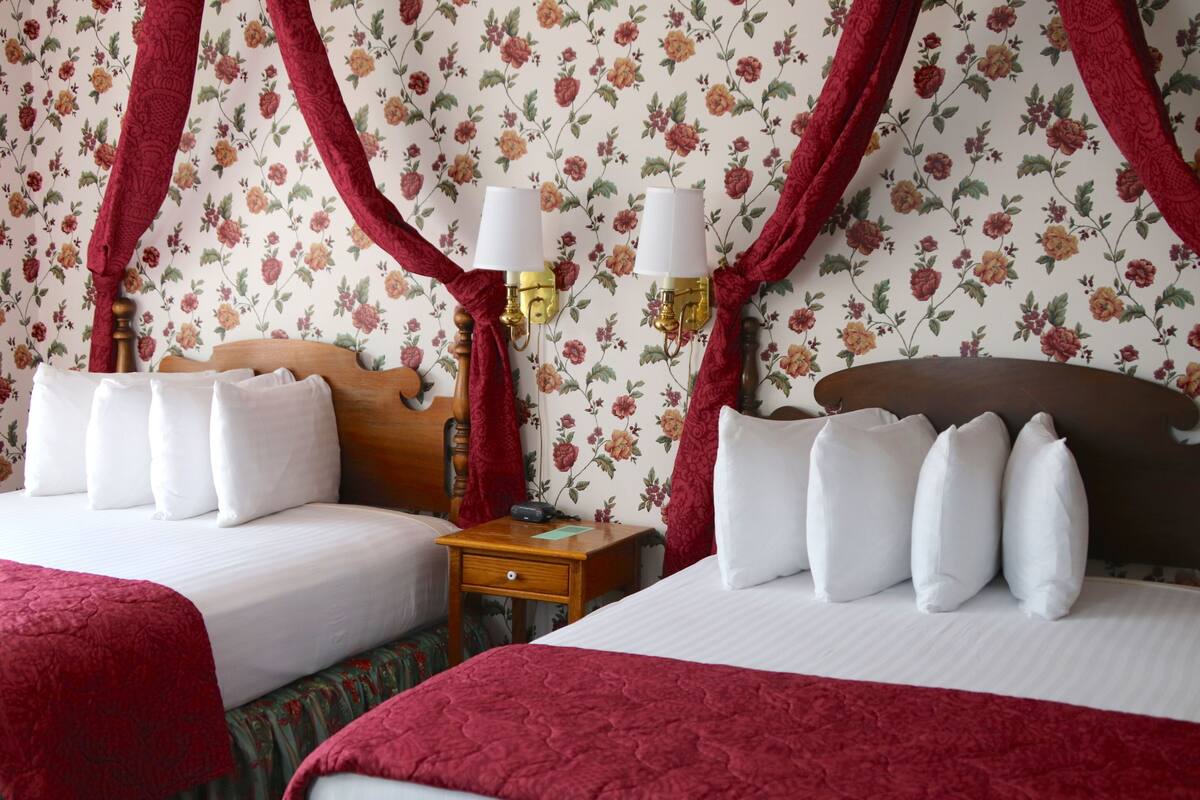 A hotel room with two beds, floral wallpaper, red curtains, and bedside lamps.