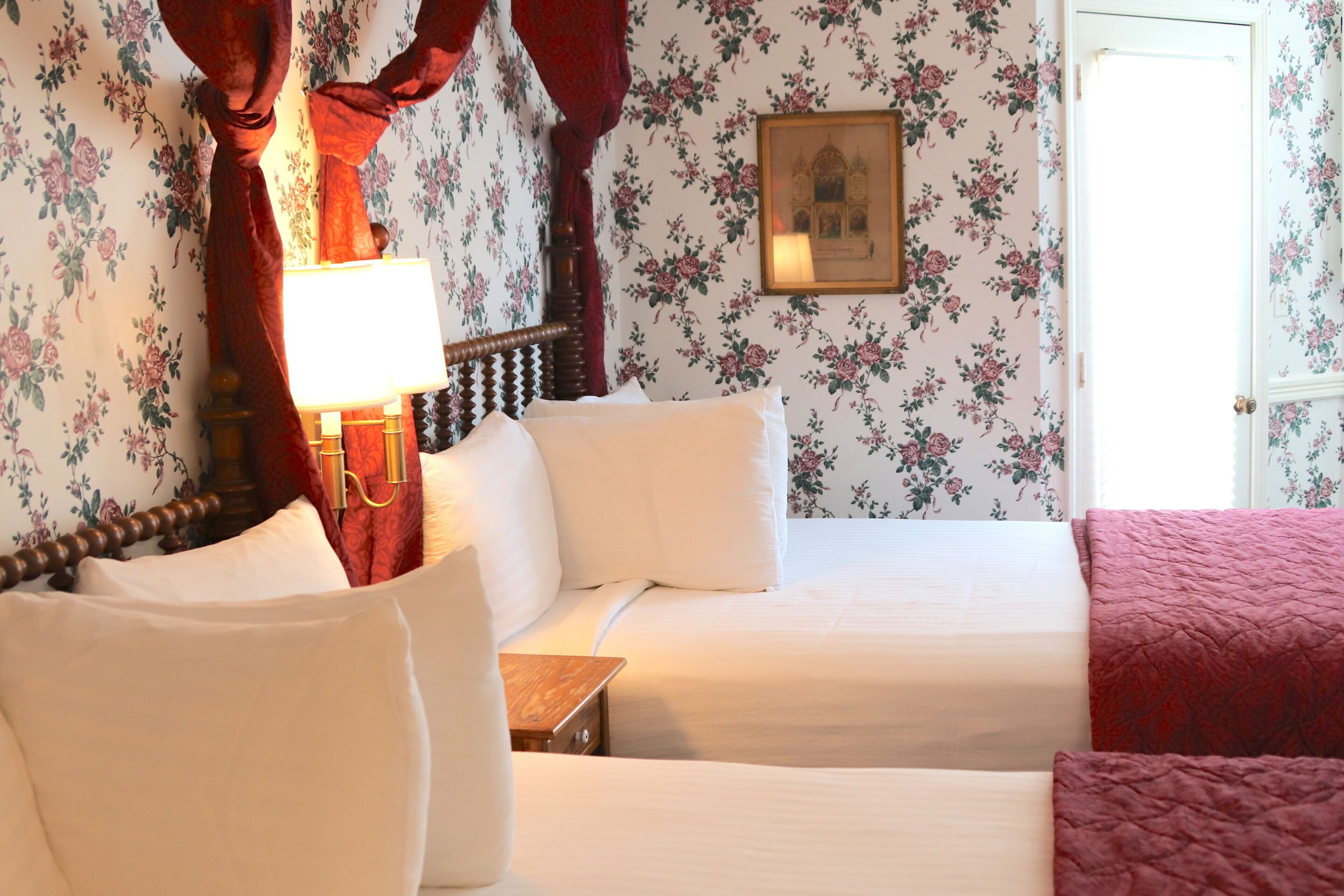 Una habitación con dos camas, cabecero de madera, cortina roja, lámpara de pared y papel tapiz con motivos florales.