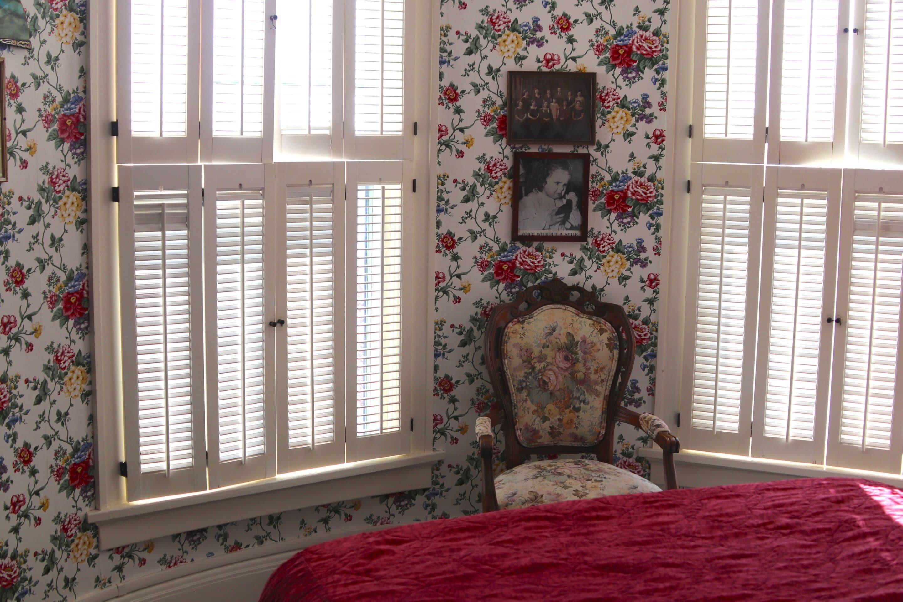 A room with floral wallpaper, a wooden chair, and a bed with a red blanket.
