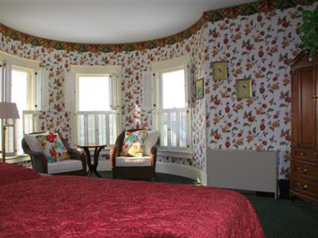 Un dormitorio con una cama grande, dos ventanas, una silla, una mesita y un aparador.