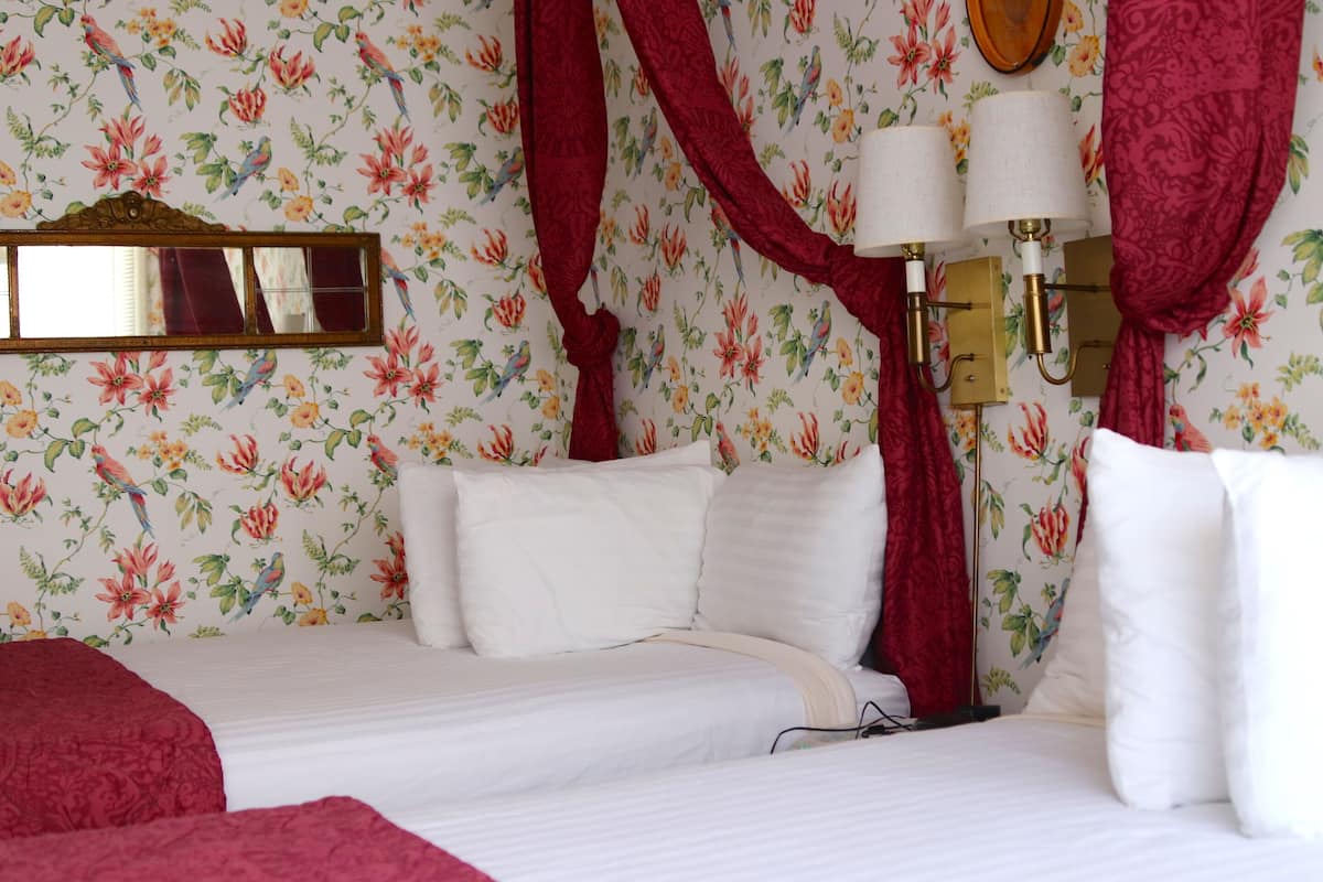 A hotel room with floral wallpaper, two beds, red curtains, a mirror, and wall-mounted lamps.