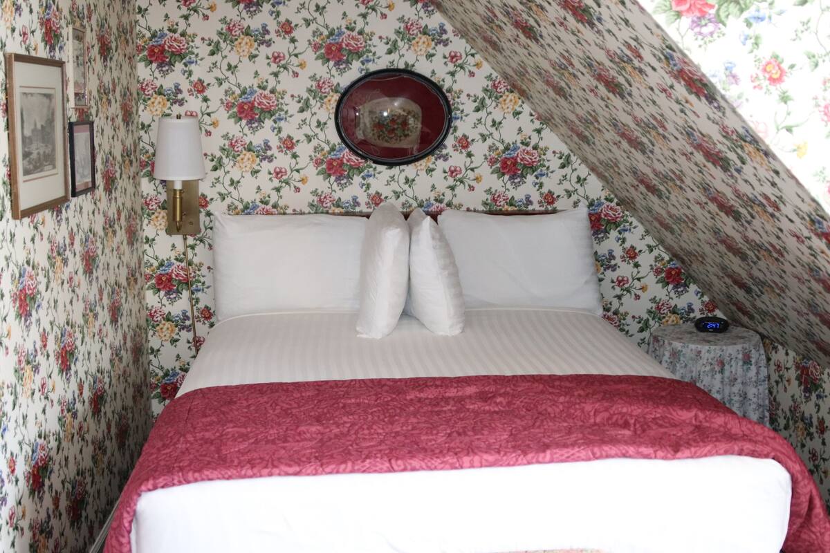 A bedroom with a floral wallpaper, a bed with white linens and a red blanket, a bedside lamp, a round mirror, and a clock on a side table.
