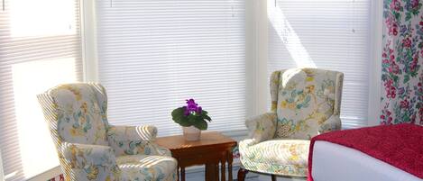 Une pièce comprenant deux fauteuils, une petite table avec un pot de fleurs et un lit recouvert d’une couverture rouge.
