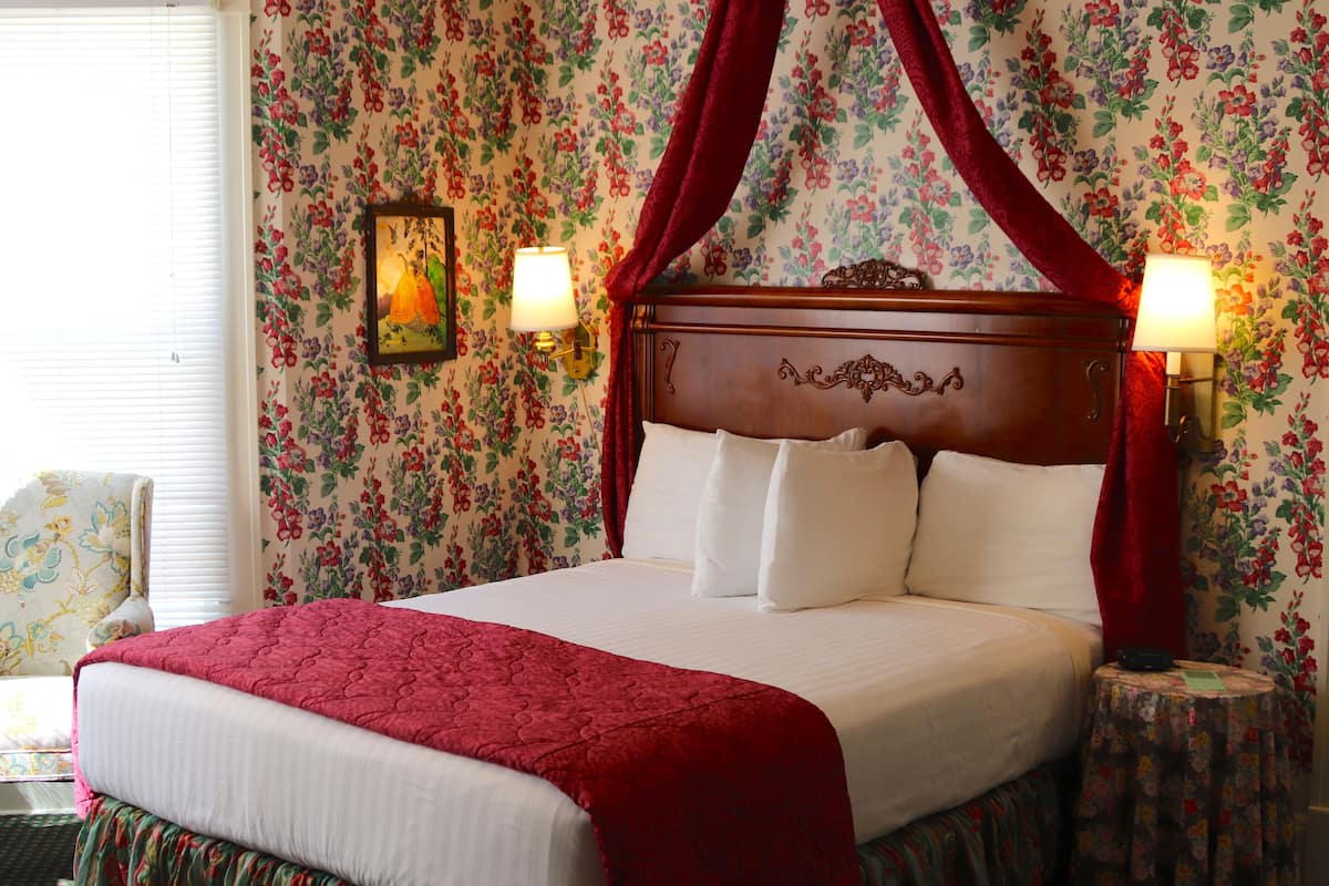 A four-poster bed with white linens, a red canopy, and floral wallpaper.