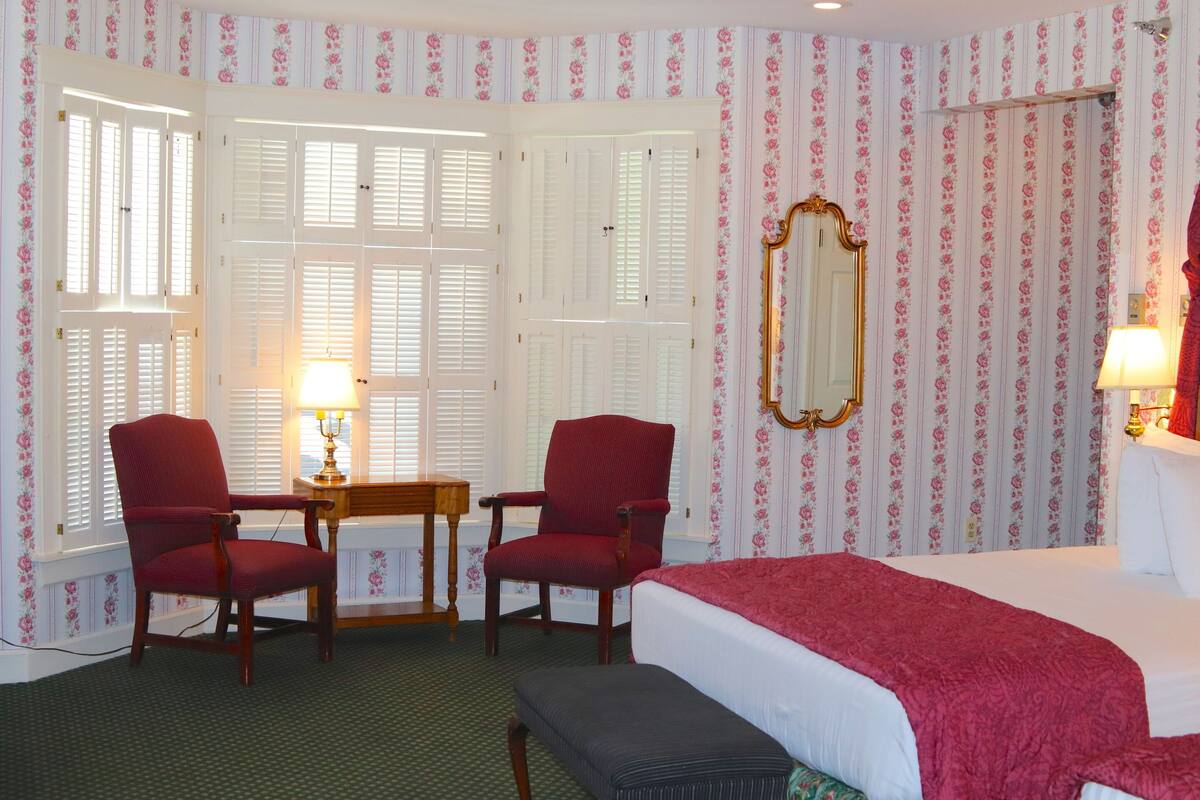 A hotel room with a bed, two armchairs, a small table with a lamp, a mirror, and floral wallpaper.