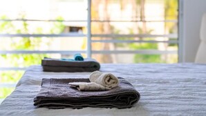 Ein Bett mit weißer Decke und zwei gefalteten Handtüchern, ein Fenster mit Jalousien und Blick auf Grün.