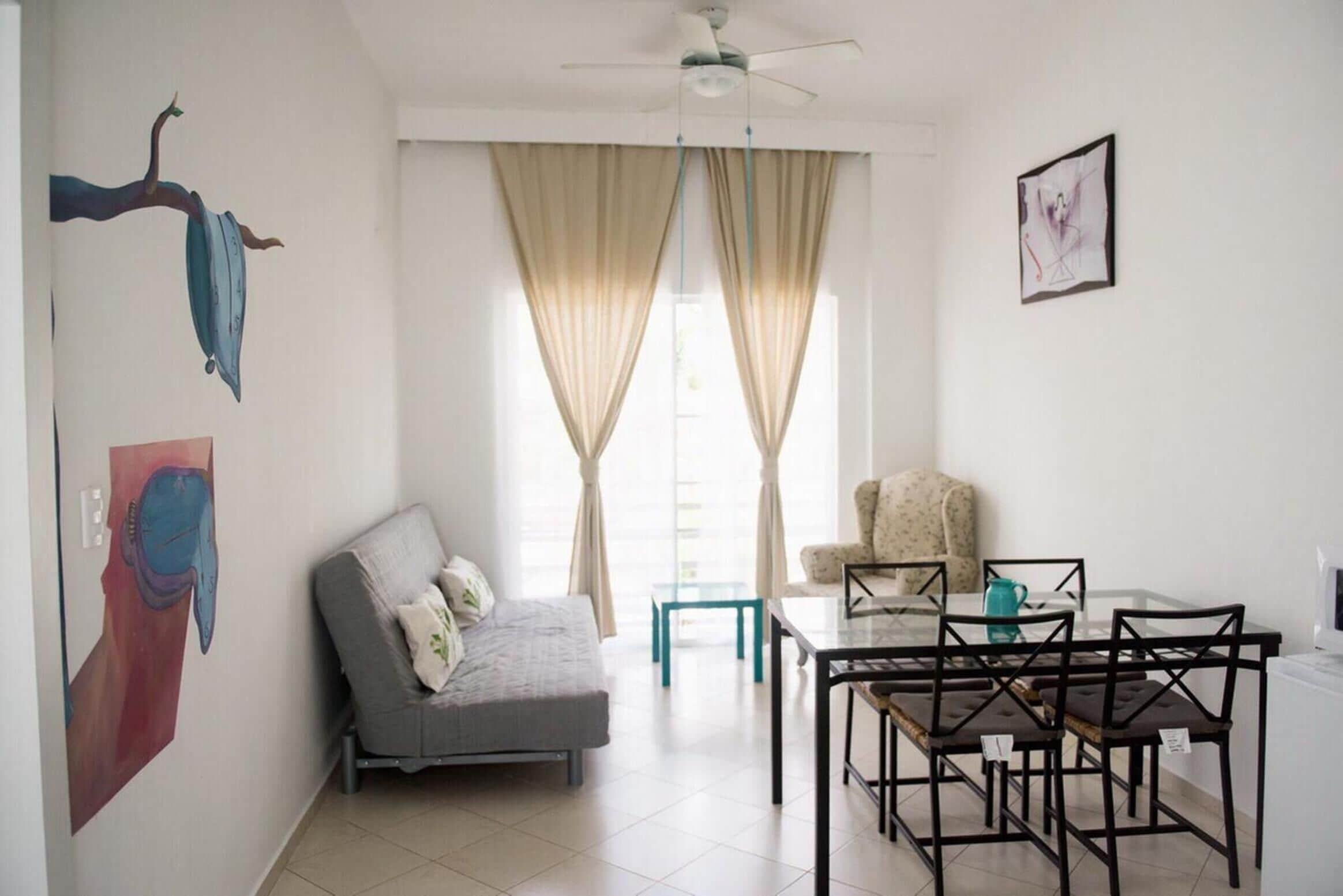 A living room with a sofa, a dining table with chairs, a ceiling fan, and large windows with curtains.