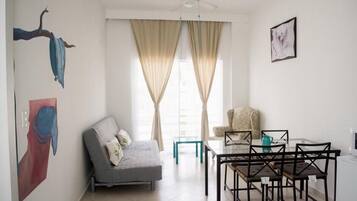A living room with a sofa, a dining table with chairs, a ceiling fan, and large windows with curtains.