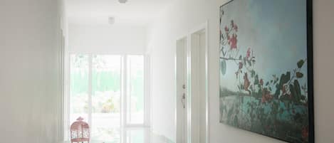Un pasillo luminoso con un ventilador de techo, una mesa de cristal con un objeto rosa, y un cuadro floral grande en la pared.