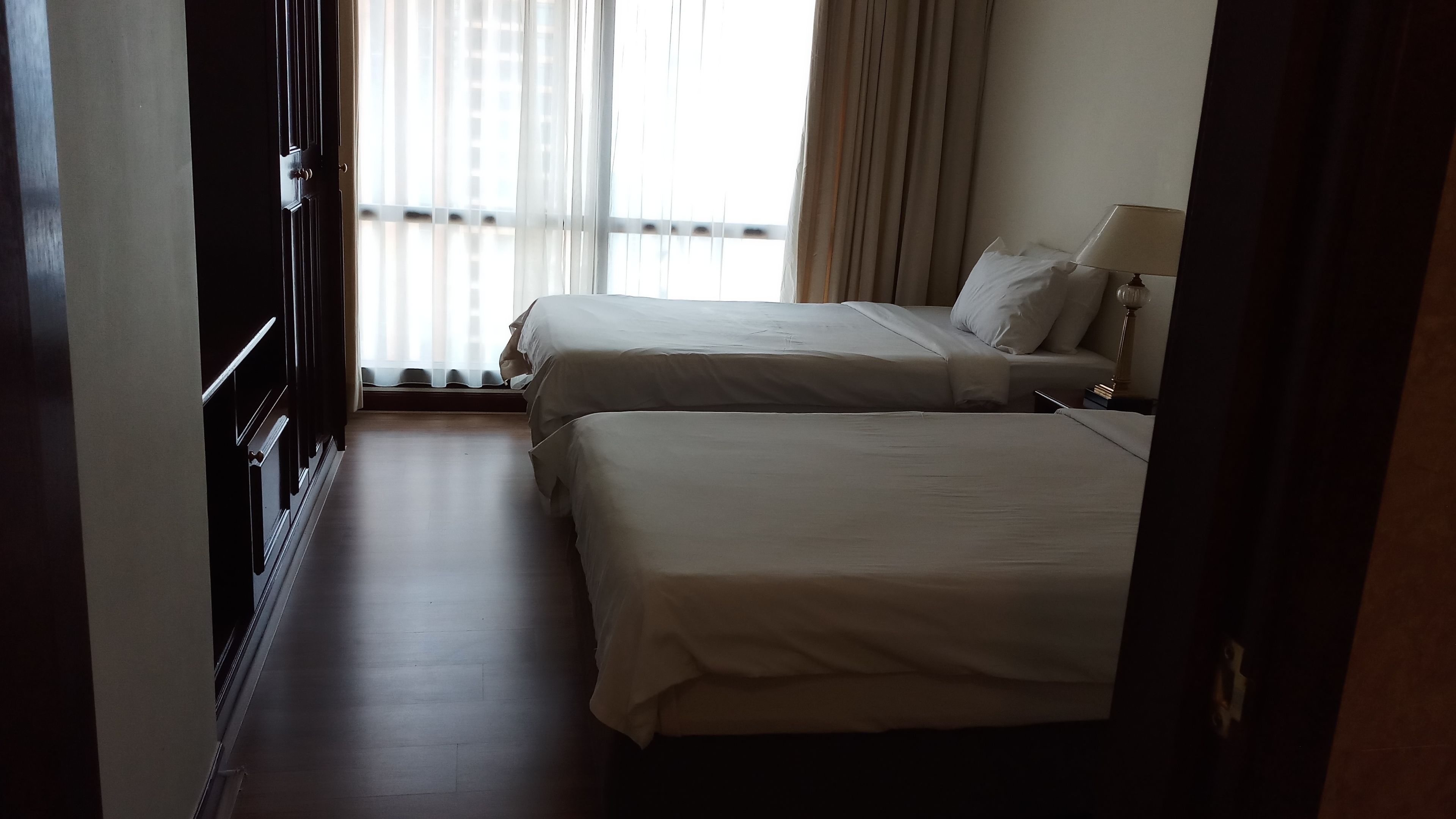 A hotel room with two beds, a window with curtains, a bedside table with a lamp, and a wooden floor.