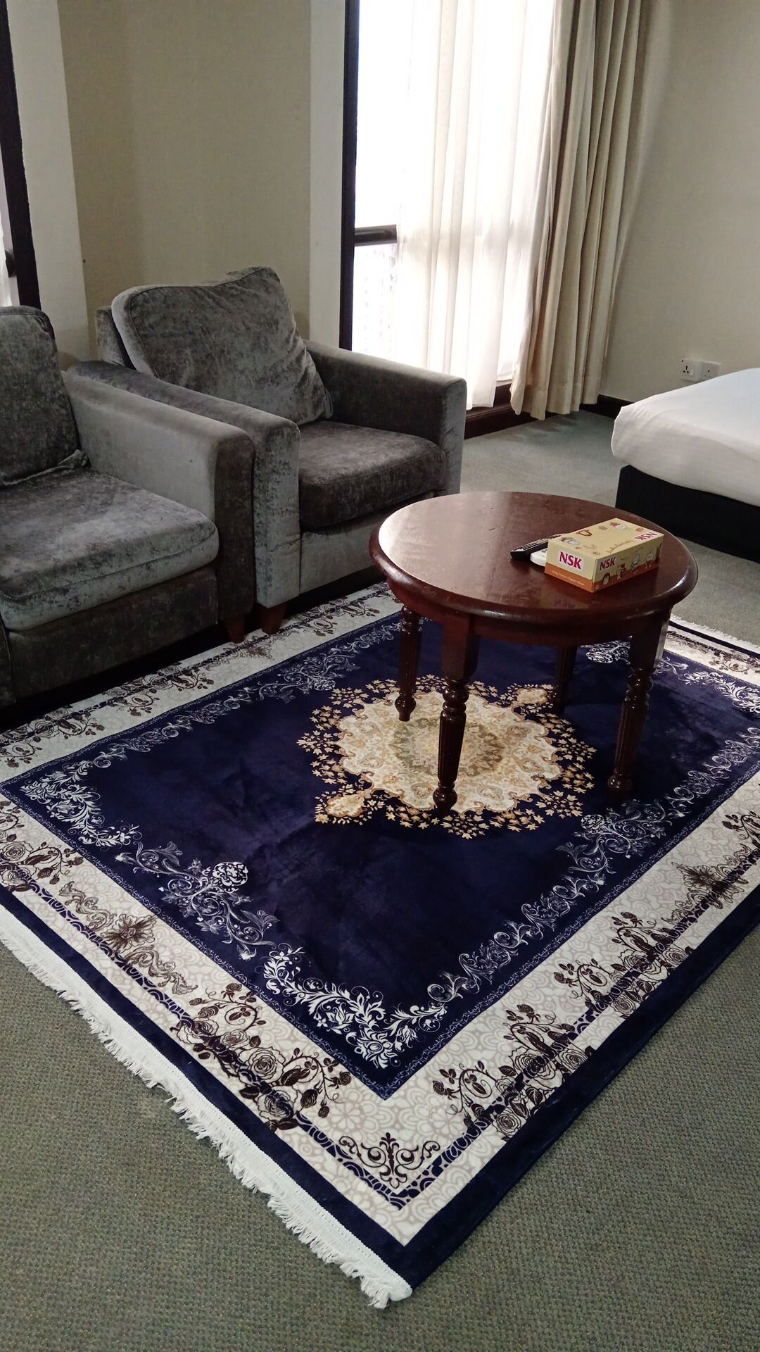 A hotel room with a grey sofa, a round wooden table with a box on it, and a large patterned rug.
