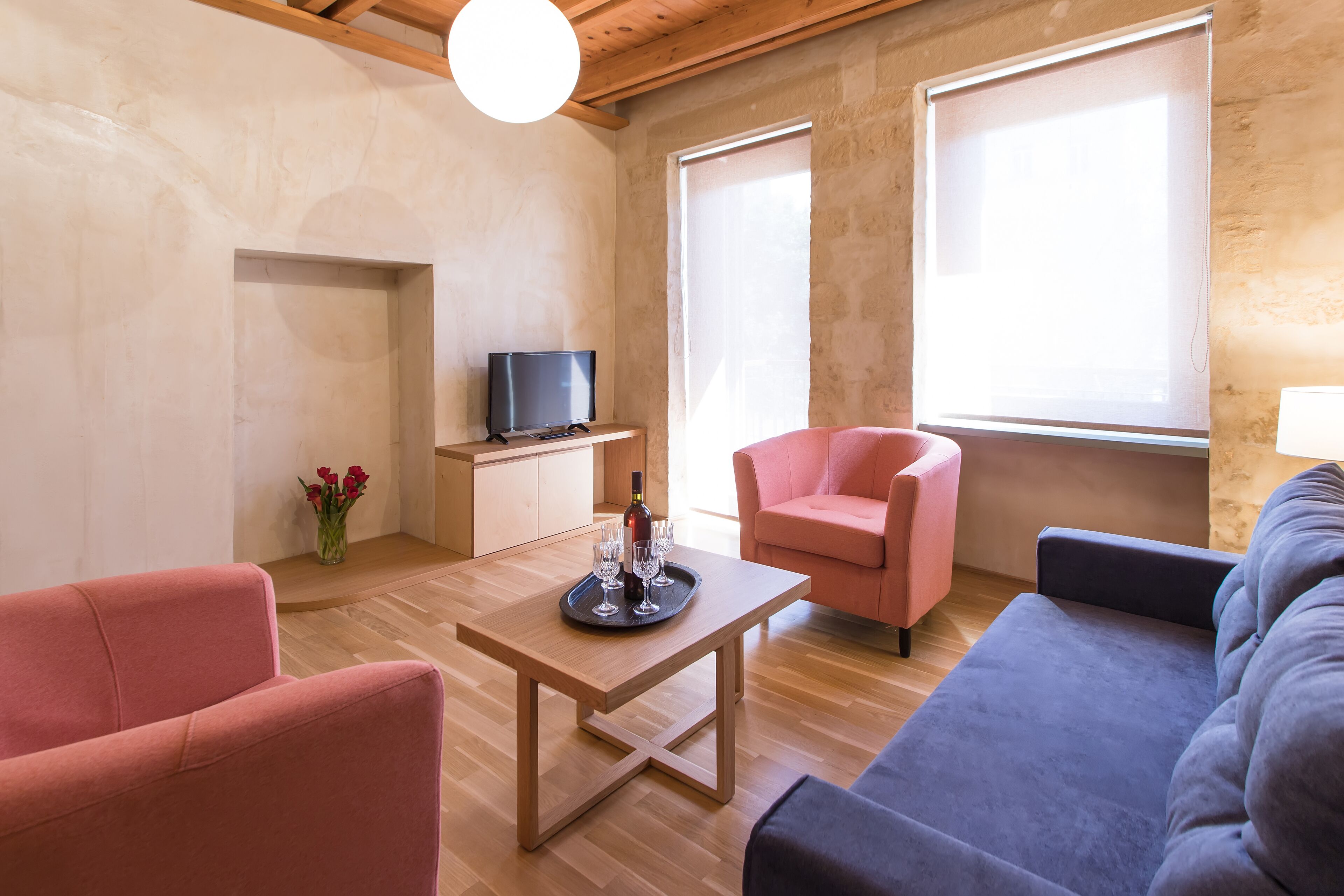 A modern living room with a wooden coffee table, a pink armchair, a blue sofa, and a television on a wooden cabinet.