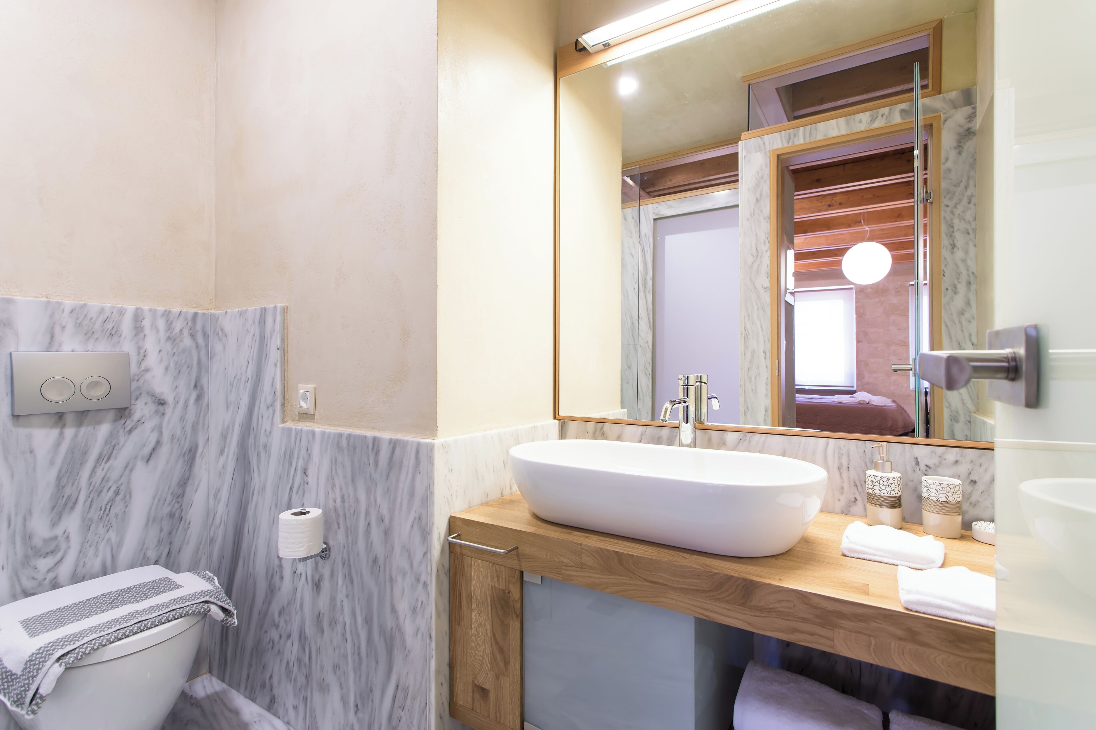 A modern bathroom with a marble wall, a wooden vanity, a large sink, and a mirror.