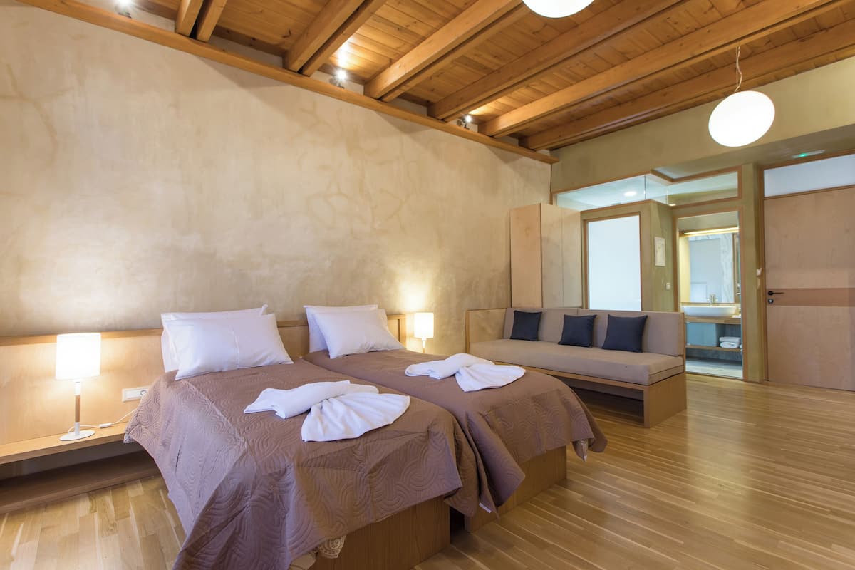 A hotel room with two beds, a sofa, wooden flooring, and a bathroom visible through an open door.