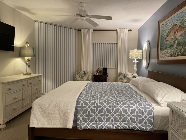 Quarto bem organizado com cama, cómoda, mesa de cabeceira, ventoinha no teto e um quadro na parede.