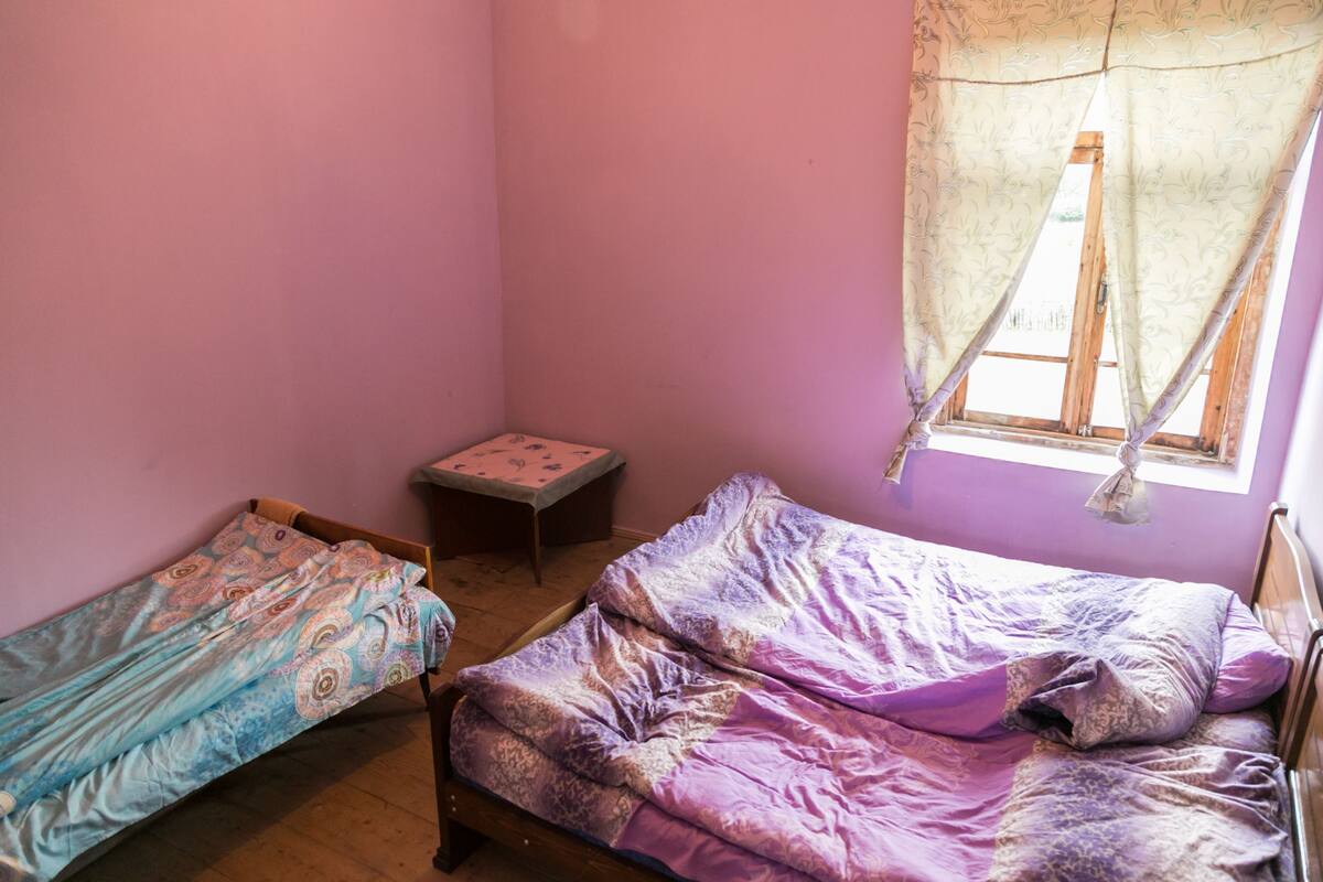 Een kleine kamer met twee eenpersoonsbedden, een tafeltje en een raam met gordijnen.