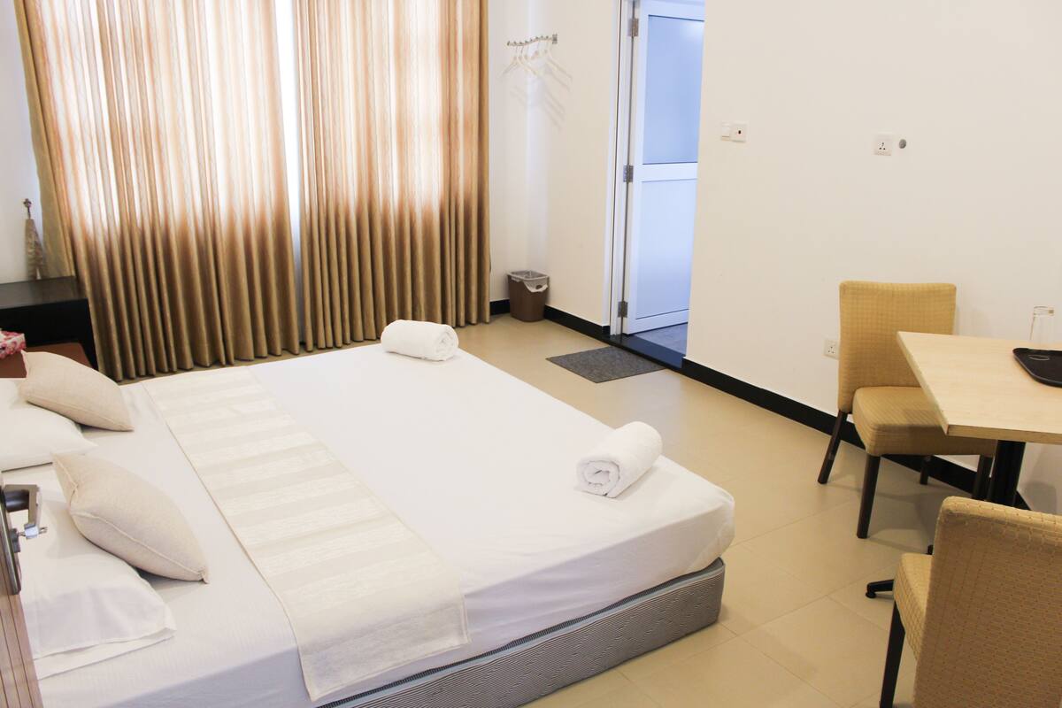 A hotel room with a bed, a desk, and a chair. There is a door and a coat rack in the background.