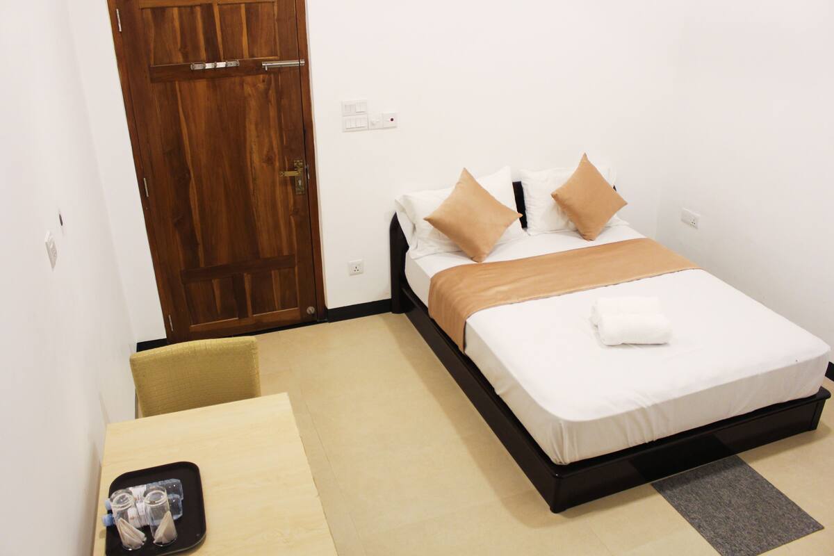 A hotel room with a bed, a wooden door, a small table, and a chair.