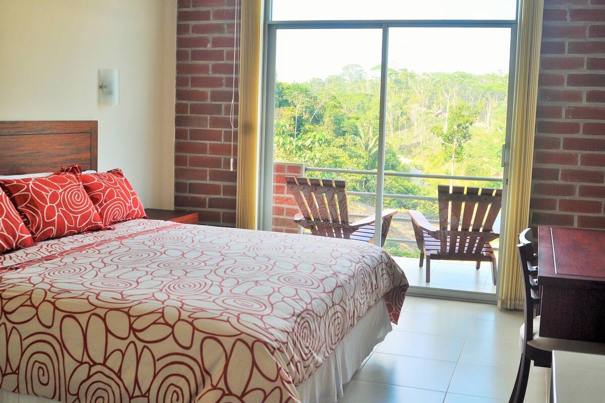 Una habitación de hotel con una cama grande, un cabecero de madera, un balcón con vistas al bosque y una mesa con sillas.