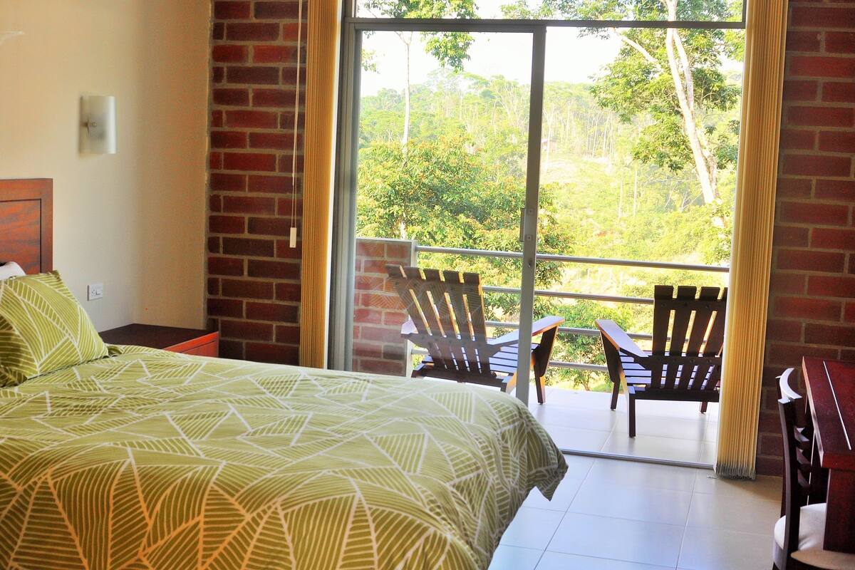 Un dormitorio con cama, una silla, una mesa y un balcón con vistas a la vegetación.