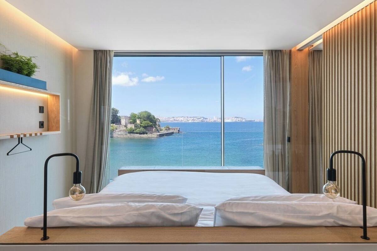 Une chambre d’hôtel avec un grand lit, deux lampes de chevet, une vue sur la mer et un paysage urbain en arrière-plan.