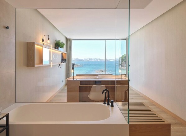 A modern bathroom with a large bathtub, a wooden vanity, and a view of the sea.