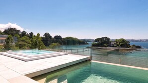 A rooftop infinity pool with clear water, surrounded by a glass railing, overlooking a coastal area with trees and buildings. - Noa Boutique Hotel (Oleiros)
