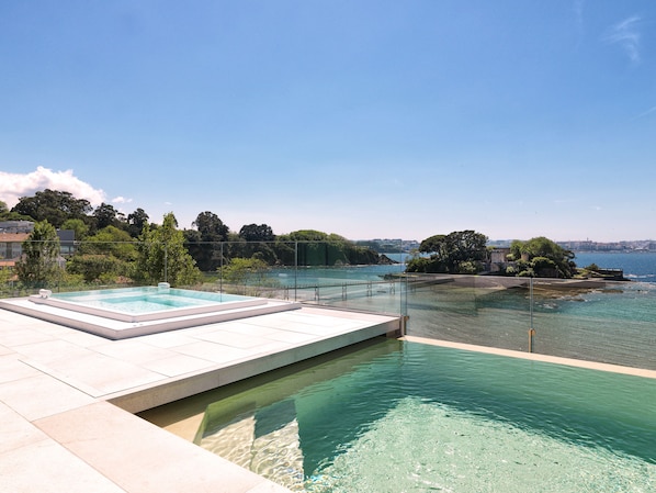A rooftop infinity pool with clear water, surrounded by a glass railing, overlooking a coastal area with trees and buildings.