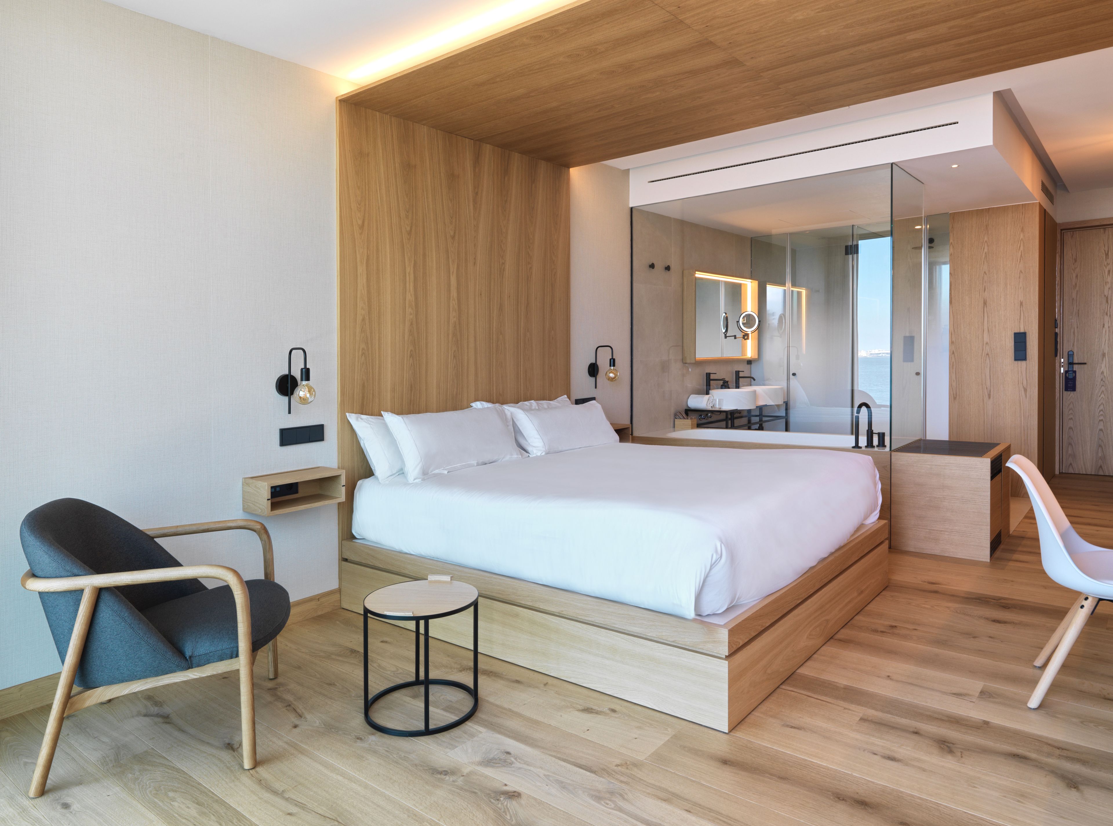 Una habitación de hotel moderna con una cama grande, una mesita auxiliar redonda, una silla, una pequeña mesa redonda, un espejo y un baño con lavabo y espejo.