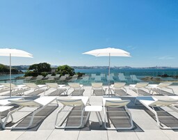Un área de piscina en la azotea con tumbonas y sombrillas, con vistas a un paisaje urbano y una masa de agua.