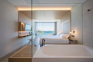 A modern hotel room with a large bed, a bathtub, a desk, and a view of the sea.