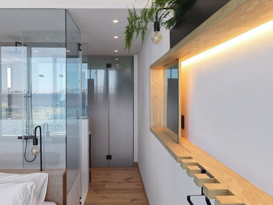 Una habitación de hotel moderna con pared de vidrio, suelo de madera, cama, banco empotrado y espejo fijado a la pared.