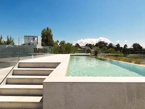 A modern outdoor swimming pool with clear blue water, surrounded by a glass fence and concrete steps leading down to the pool.