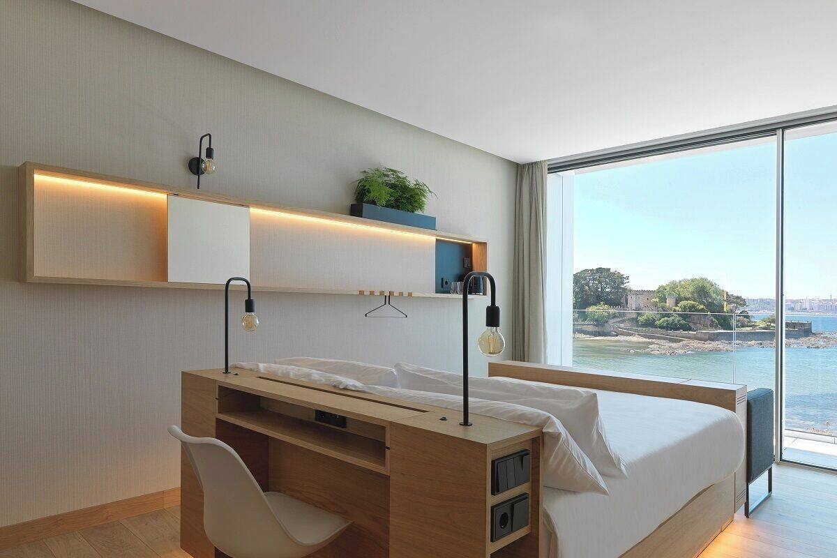 Une chambre moderne avec un lit, un bureau et une chaise, une grande fenêtre donnant sur la mer, et des étagères intégrées ornées de plantes.