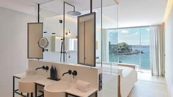 Un baño moderno con tocador doble lavamanos, un espejo grande y una ducha. La habitación cuenta con un ventanal con vistas a una masa de agua y una isla distante.