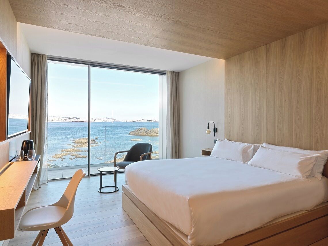 Una habitación de hotel moderna con una cama grande, un escritorio y una silla. La habitación tiene un ventanal con vistas al mar.