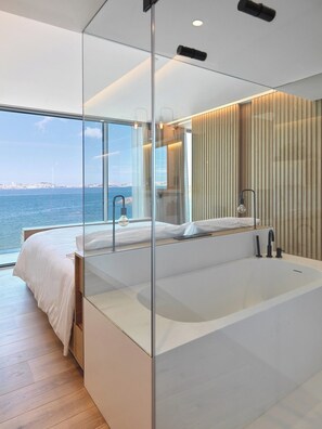 A modern bedroom with a large bed, a bathtub, and a view of the sea.