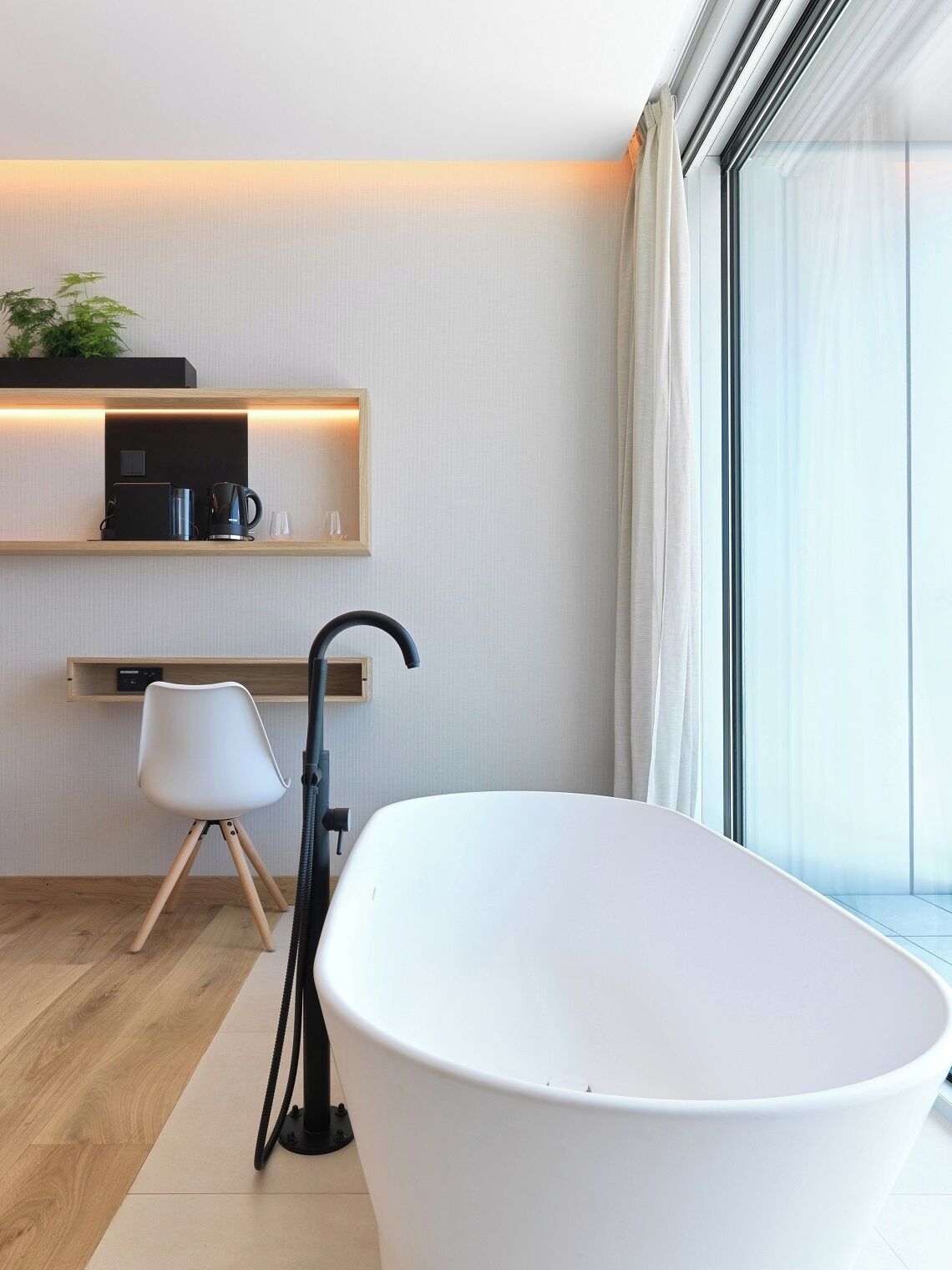 Une salle de bain moderne avec une baignoire sur pieds, une petite étagère avec une machine à café, une chaise et une grande fenêtre avec des rideaux.