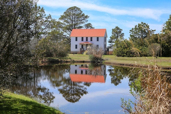 A two-story house with a red roof, surrounded by a pond and trees.