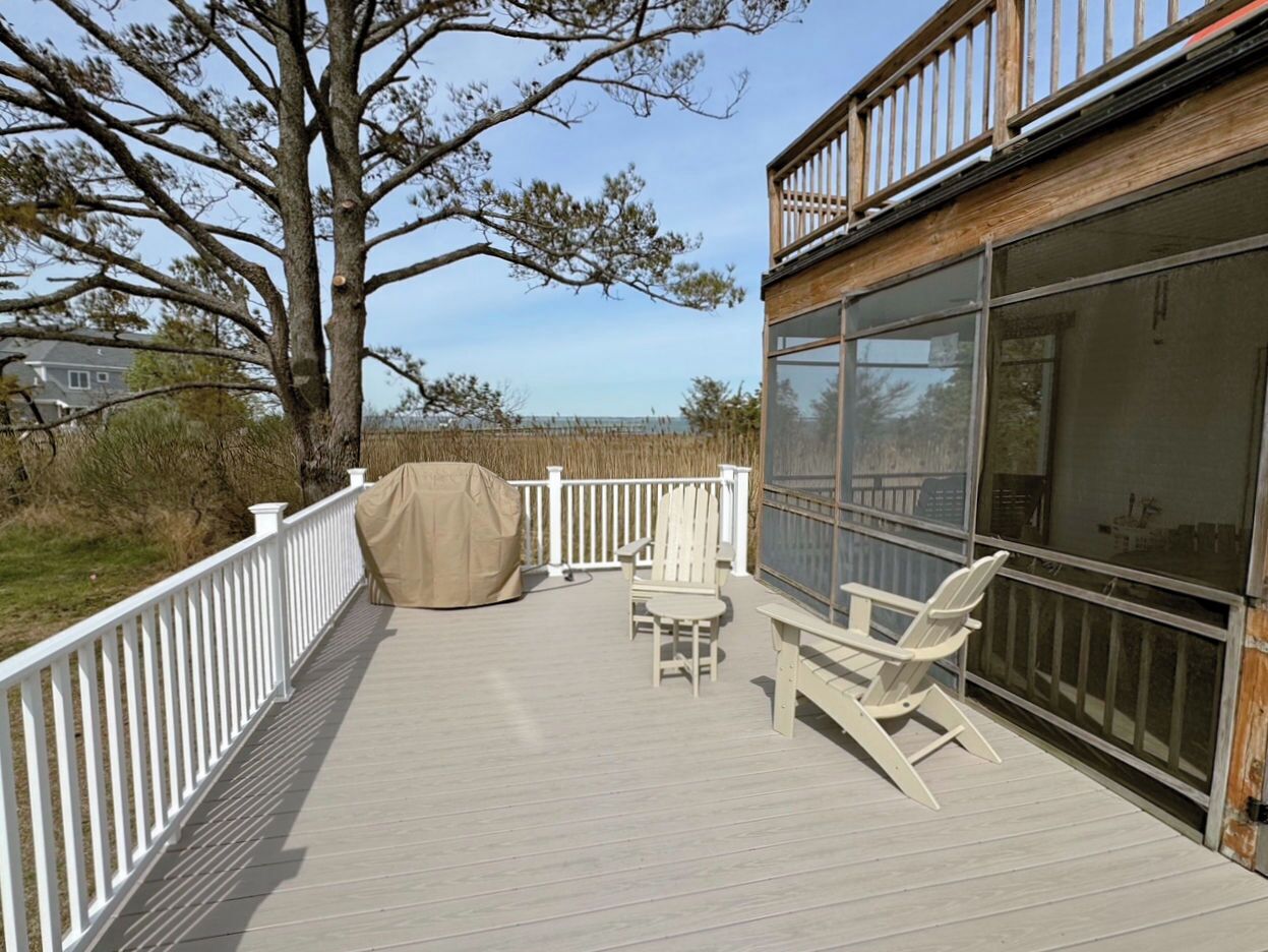 A deck with a white railing, two Adirondack chairs, a covered table, and a large window overlooking a natural landscape.