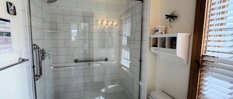 Et bruseafsnit med glasvægge, et hvidt toilet og et vindue med persienner.